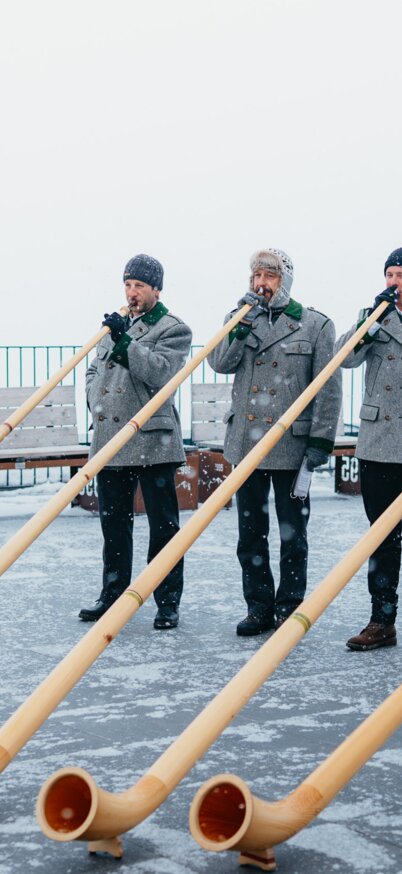 Fünf Alphornbläser spielen auf der verschneiten Aussichtsplattform am Dachstein Gletscher vor winterlicher Bergkulisse. | © Stefan Simonovic, studio draussen