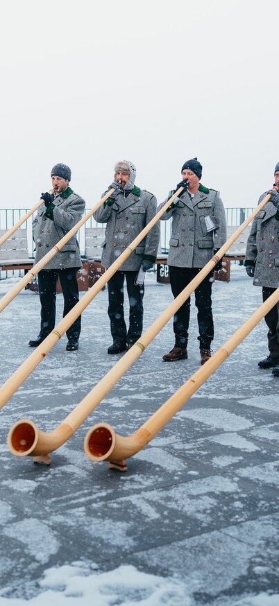 Fünf Alphornbläser spielen auf der verschneiten Aussichtsplattform am Dachstein Gletscher vor winterlicher Bergkulisse. | © Stefan Simonovic, studio draussen