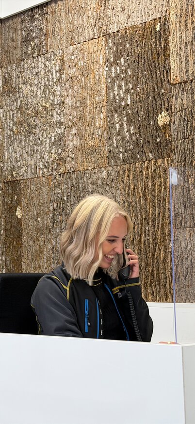 Mitarbeiterin im Info- und Servicebereich des Hauser Kaibling sitzt an einem modernen Empfangstresen und telefoniert, vor einer Wand mit Naturholzoptik. | © Hauser Kaibling