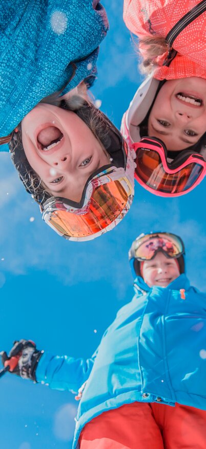 Vier Kinder und eine erwachsene Person blicken lachend in die Kamera. Die Aufnahme entstand aus der Froschperspektive auf einer verschneiten Skipiste in Ski amadé. | © Claudia Ziegler
