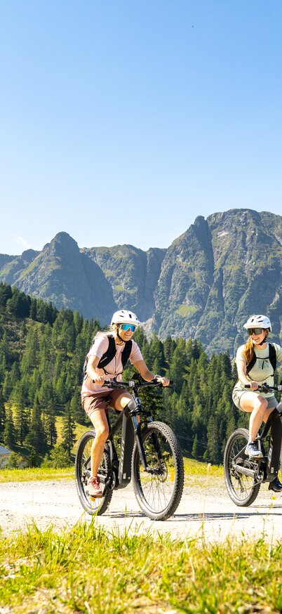 Zwei Radfahrerinnen auf E-Mountainbikes lächeln während der Fahrt auf einen breiten Weg am Hauser Kaibling entlang, im Hintergrund eine beeindruckende Bergkulisse. | © Josh Absenger