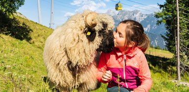 A child is sitting in a mountain meadow and giving a sheep a kiss. In the background, you can see a gondola, the mountain landscape, and a sunny, blue sky. | © Josh Absenger