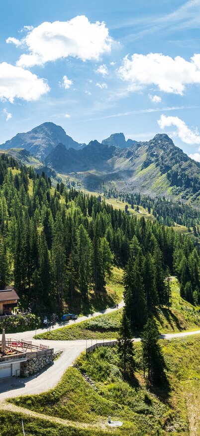 Die Genussalm am Hauser Kaibling mit Sonnenterrasse, eingebettet in eine grüne Berglandschaft mit Nadelwäldern und hohen Gipfeln. | © Josh Absenger