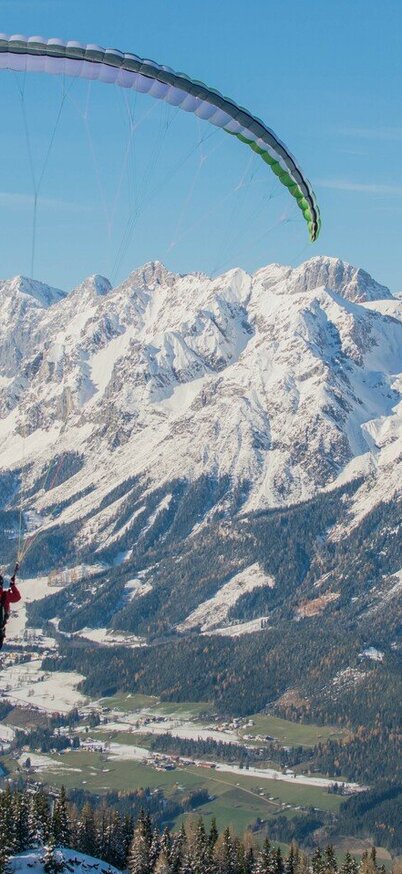 Paragleiter im Winter über den verschneiten Hängen des Hauser Kaibling mit Blick auf das Dachsteinmassiv und das Ennstal. | © Hauser Kaibling