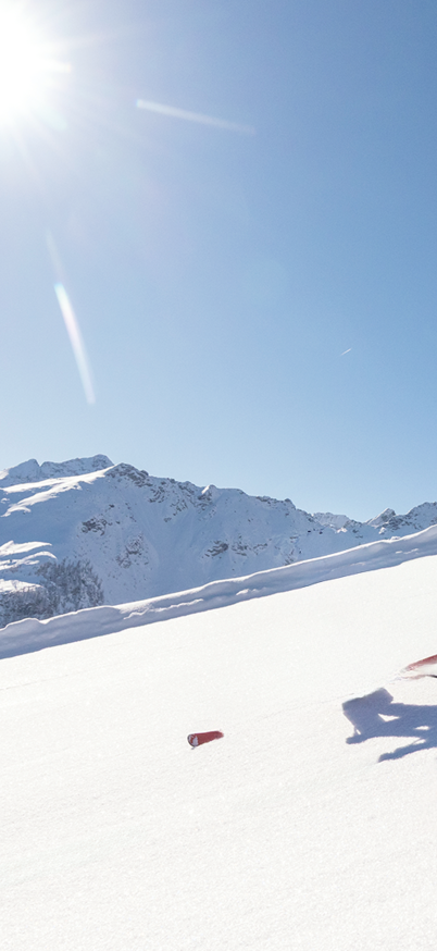 Zwei Skifahrerinnen carven in farbenfroher Skibekleidung bei strahlendem Sonnenschein die frisch präparierte Piste am Hauser Kaibling hinunter, umgeben von tief verschneiten Bergen. | © Bernhard Moser