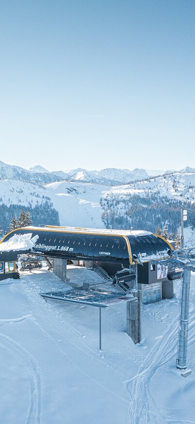 Panoramablick auf die Bergstation der Kaiblinggrat 8er-Sesselbahn am Hauser Kaibling mit markantem Sendeturm, verschneiter Skipiste und traumhafter Winterkulisse - im Hintergrund das Dachsteinmassiv. | © Josh Absenger