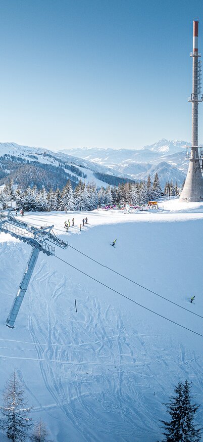 Panoramablick auf die Bergstation der Kaiblinggrat 8er-Sesselbahn am Hauser Kaibling mit markantem Sendeturm, verschneiter Skipiste und traumhafter Winterkulisse - im Hintergrund das Dachsteinmassiv. | © Josh Absenger