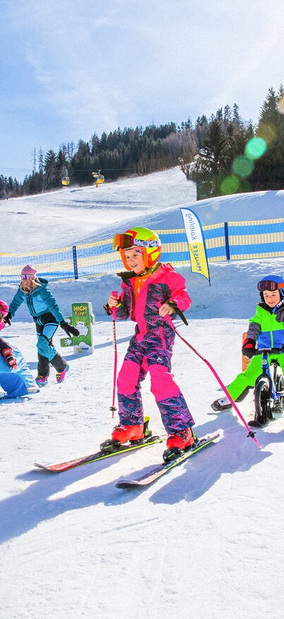 Skifahren für Kinder und Anfänger im Wollis Kids Park am Hauser Kaibling | © René Eduard Perhab Kinder genießen einen sonnigen Wintertag im Wollis Kids Park am Hauser Kaibling – ein Mädchen fährt mit roten Skiern die Piste hinunter, während ein Junge daneben mit einem Kinderlaufrad über den Schnee rollt. Im Hintergrund spielen weitere Kinder unter strahlend blauem Himmel. | © René Eduard Perhab