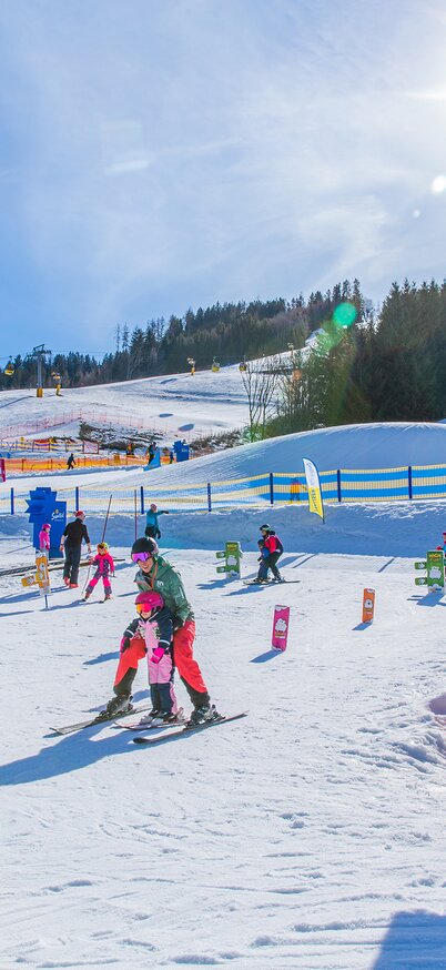 Children ride up the slope on the "Lockis Zauberteppich" magic carpet lift in the sunny Wollis Kids Park, surrounded by colorful gates and figures. | © René Eduard Perhab