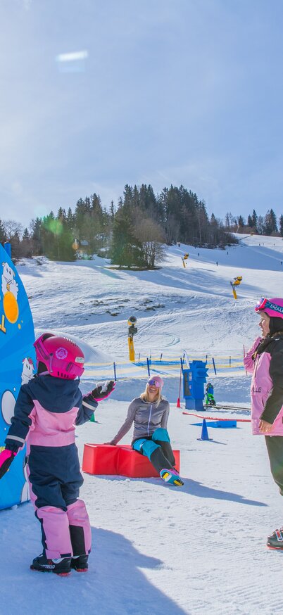 Kinder werfen lachend Schneebälle durch die bunten Öffnungen einer Spielwand im Wollis Kids Park, während die Wintersonne über den verschneiten Hängen scheint. | © René Eduard Perhab