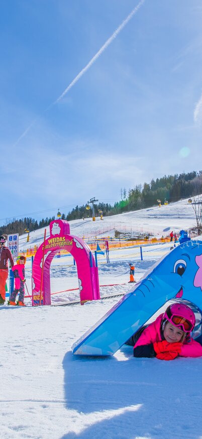 Drei Kinder spielen im sonnigen Wollis Kids Park am Hauser Kaibling – zwei sitzen lachend auf einem blauen Elefantenspielgerät, während ein weiteres Kind darunter hindurchkriecht. | © René Eduard Perhab
