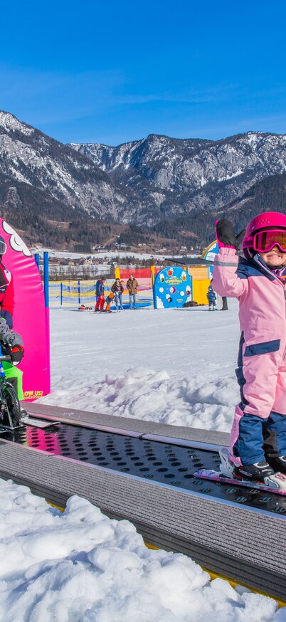 Kinder fahren auf dem pinken „Weidas Zauberteppich“ im Wollis Kids Park am Hauser Kaibling den Förderbandlift hinauf. Ein Mädchen im rosa Skianzug winkt lachend in die Kamera, während hinter ihr weitere Kinder mit Ski und Laufrädern begleitet von einer Erwachsenen den Zauberteppich nutzen. | © René Eduard Perhab