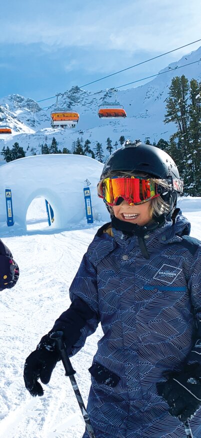 Three children in colorful ski suits stand laughing on the XXL Funslope, while a large snow tunnel, orange chairlifts and the snow-covered mountains under a blue sky can be seen in the background. | © Josh Absenger