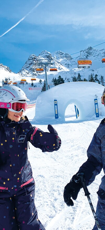 Three children in colorful ski suits stand laughing on the XXL Funslope, while a large snow tunnel, orange chairlifts and the snow-covered mountains under a blue sky can be seen in the background. | © Josh Absenger
