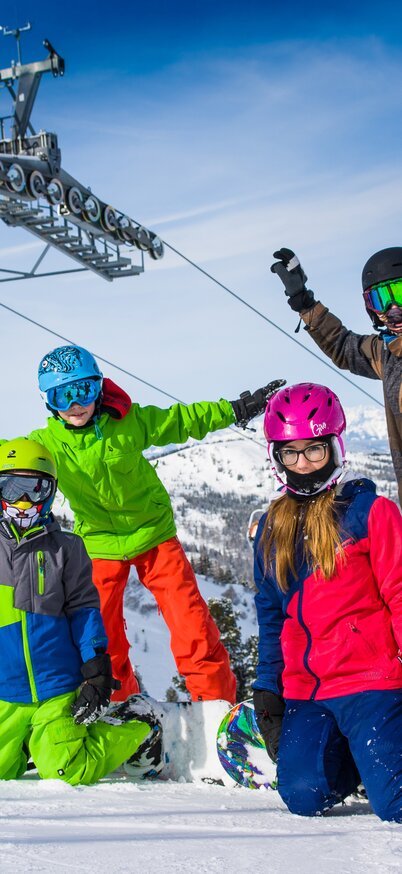 Four children in colorful ski and snowboard equipment pose laughing in the snow at Hauser Kaibling, while a chairlift with orange weather protection hoods passes behind them. | © Foto Atelier Wolkersdorfer