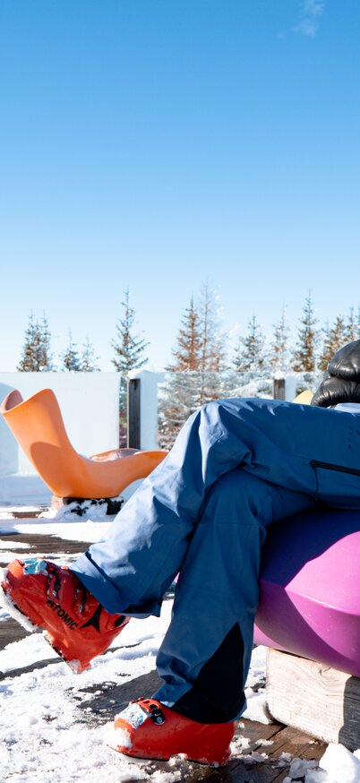 A skier sits relaxed on a purple armchair on the Genussinsel at Hauser Kaibling, surrounded by other colorful armchairs under a blue sky. | © Josh Absenger