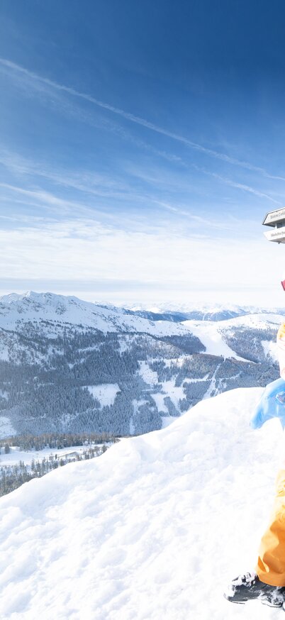 Zwei Skifahrerinnen lachend am Gipfelkreuz des Hauser Kaibling, die Skier im Arm, umgeben von einem Panorama schneebedeckter Berge unter blauem Himmel. | © Bernhard Moser