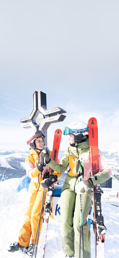 Zwei Skifahrerinnen lachend am Gipfelkreuz des Hauser Kaibling, die Skier im Arm, umgeben von einem Panorama schneebedeckter Berge unter blauem Himmel. | © Bernhard Moser 
