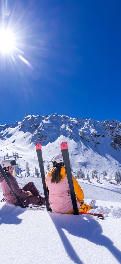 Zwei Skifahrerinnen sitzen entspannt im Schnee am Hauser Kaibling, lehnen sich an ihre Skier und genießen bei strahlendem Sonnenschein und wolkenlosem Himmel die Aussicht auf die verschneiten Berge. | © Bernhard Moser