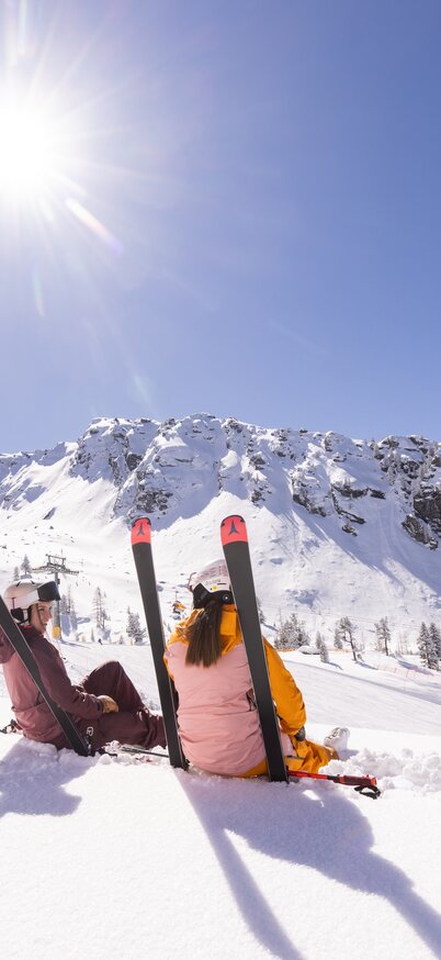 Two skiers sit relaxed in the snow at Hauser Kaibling, leaning on their skis and enjoying the view of the snow-covered mountains in bright sunshine and a cloudless sky. | © Bernhard Moser