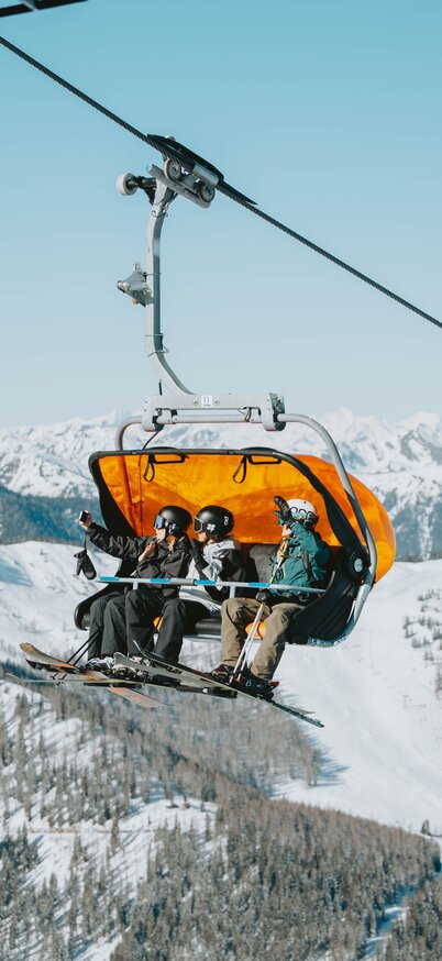 Three skiers sit on a chairlift with an orange weather protection hood and ride over the snow-covered slopes of Hauser Kaibling, with an impressive mountain panorama in the background. | © Robin Betzel
