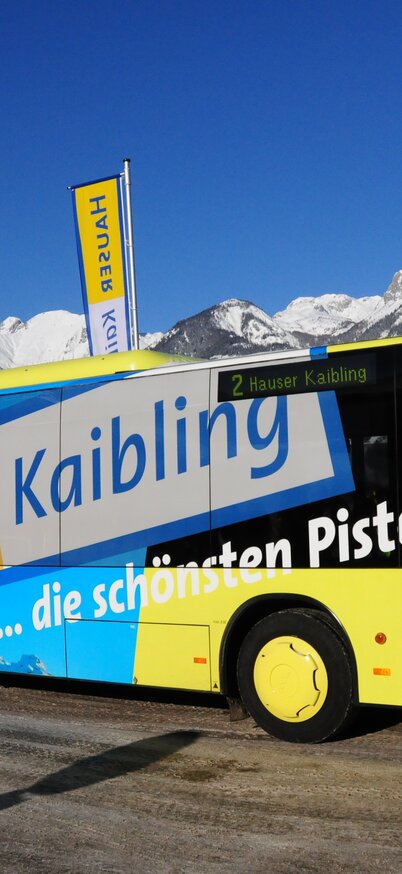 Skibus mit auffälliger Hauser-Kaibling-Beschriftung und Slogan „… die schönsten Pisten“ vor verschneiter Bergkulisse. | © Hauser Kaibling