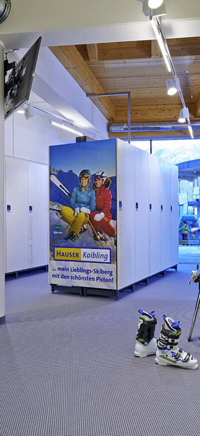 Ski depot at Hauser Kaibling with white lockers, ski boot storage and a poster with winter sports enthusiasts. | © Andy Sillaber