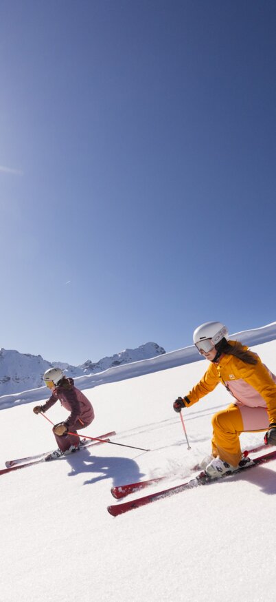 Zwei Skifahrerinnen carven in farbenfroher Skibekleidung bei strahlendem Sonnenschein die frisch präparierte Piste am Hauser Kaibling hinunter, umgeben von tief verschneiten Bergen. | © Bernhard Moser