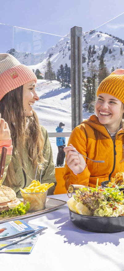 Two women eating burgers, chips and salad on the sun terrace of the Kaiblingalm at Hauser Kaibling, with the ski slope and snow-covered peaks in the background. | © Bernhard Moser