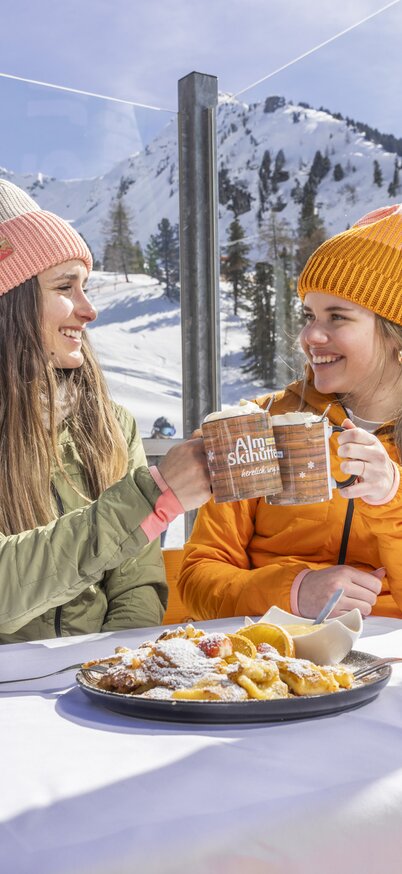 Zwei junge Frauen stoßen auf der Sonnenterrasse der Kaiblingalm am Hauser Kaibling mit Kakao an, vor ihnen steht ein Teller mit Kaiserschmarrn, im Hintergrund schneebedeckte Berge. | © Bernhard Moser