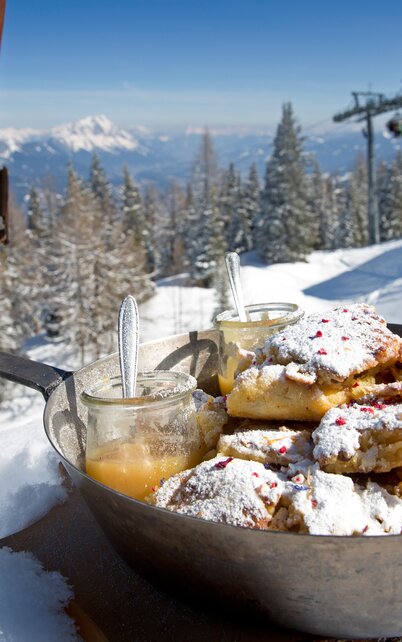 Frischer Kaiserschmarrn mit Staubzucker und Apfelmus serviert in einer Pfanne im Schnee, mit Blick auf die Pisten und Berge am Hauser Kaibling. Dahinter ein altes Paar Holzski. | © Herbert Raffalt