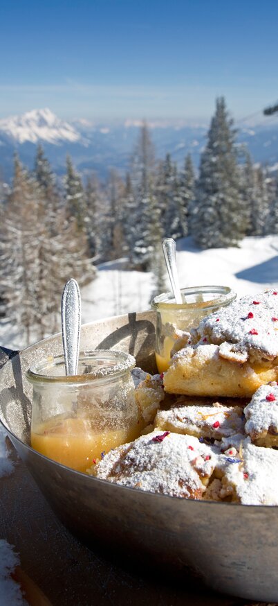 Frischer Kaiserschmarrn mit Staubzucker und Apfelmus serviert in einer Pfanne im Schnee, mit Blick auf die Pisten und Berge am Hauser Kaibling. Dahinter ein altes Paar Holzski. | © Herbert Raffalt