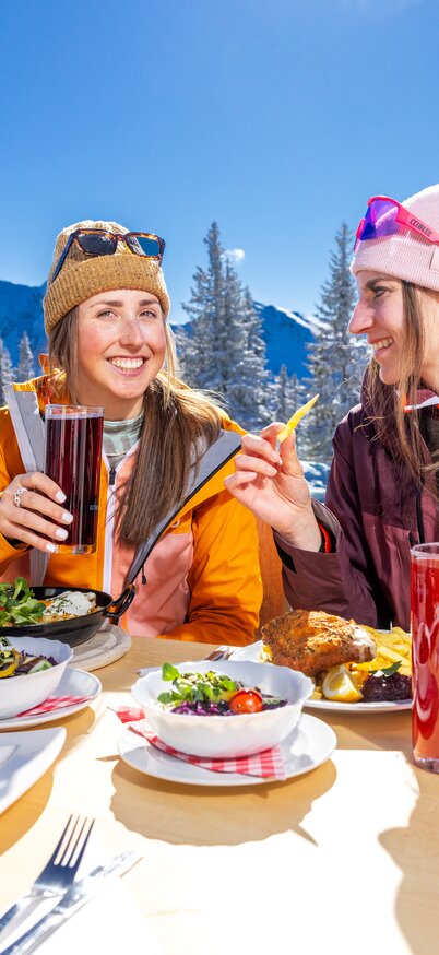Zwei Frauen sitzen auf der sonnigen Hüttenterrasse des TOMiziel am Hauser Kaibling, stoßen an und genießen regionale Schmankerl. | © Bernhard Moser
