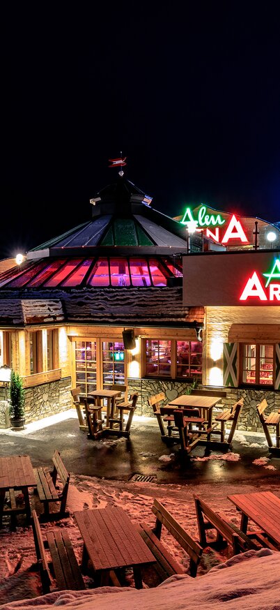 Im Bild eine Außenansicht der AlmArenA, an der Talstation der Hauser Kaibling Seilbahn, bei Nacht, festlich beleuchtet mit einladender Terrasse und Holztischen. | © Martin Huber