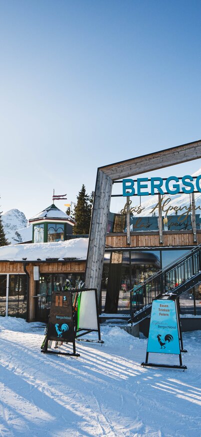 Vorderansicht des Bergschlössl Hauser Kaibling mit markantem Holzrahmen und Schriftzug, umgeben von Skifahrern und klarer Wintersonne. | © Robin Betzel