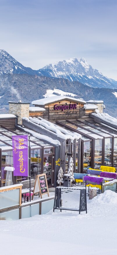 Außenansicht der Gumpenbar am Hauser Kaibling mit Glasfront, Panoramaterrasse und Blick auf die verschneiten Berge. | © Harald Steiner Fotografie