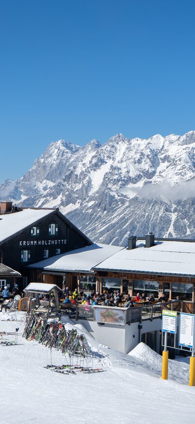 Exterior view of the Krummholzhütte at Hauser Kaibling with skiers and snow-covered mountains in the background. | © Josh Absenger