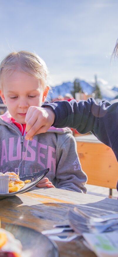 Zwei Kinder essen süße Nachspeisen und Kuchen auf der Sonnenterrasse der Krummholzhütte. | © René Eduard Perhab