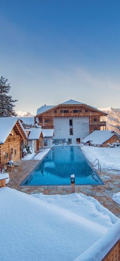 Beheizter Außenpool im Natur- und Wellnesshotel Höflehner am Hauser Kaibling, umgeben von verschneiten Chalets und Bergpanorama. | © Rene Strasser