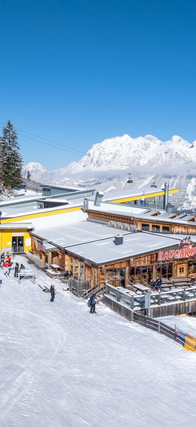 Skifahrer genießen die Sonnenterrasse der Stoni´s Rauchkuchl mit Blick auf die verschneiten Berge. Daneben die Gondelbahn und weitläufige Skipisten. | © Josh Absenger