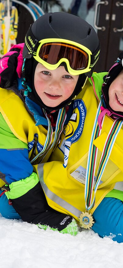 Zwei Kinder in bunter Skikleidung mit gelben Westen und Helmen sitzen lachend im Schnee, stolz mit Medaillen um den Hals nach einem Skikurs am Hauser Kaibling. | © Patrick Putre