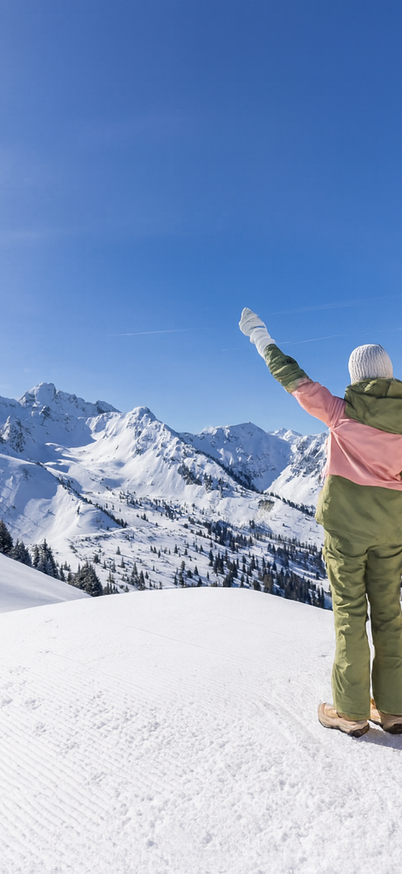 Two people in colorful winter clothing stand on a snowy mountain peak with their arms raised and look out over the surrounding Alps. A bright blue sky and snow-covered mountains stretch as far as the horizon. | © Original photo Bernhard Moser | edited with KI/Photoshop