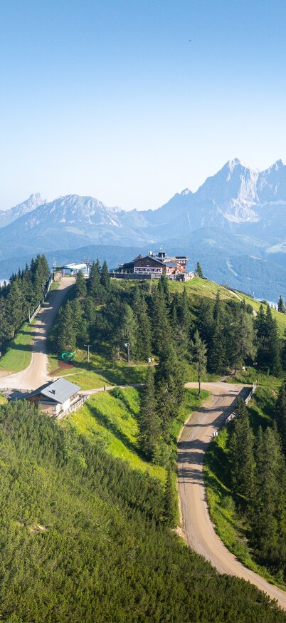 Panoramablick vom Hochwurzen-Gipfel Richtung Dachstein | © Josh Absenger Grüne Hochwurzen-Gipfelkulisse mit Hütte, Bergstraße und Blick auf das Ennstal und das Dachsteinmassiv | © Josh Absenger