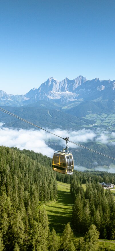 The Hochwurzen summit lift ascends with a view of the Dachstein massif and clouds in the valley. | © Josh Absenger