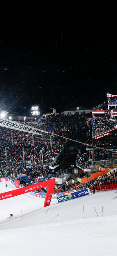 © Martin Huber Nightrace 2024 auf der Planai, Slalom, Herren, 1. Durchgang, im Bild ein Tiefblick in den Ziel-Bereich mit Skygate, Tribünen, Hohenhaus Tenne und Yellow-Tribüne sowie Tommaso Sala (ITA) im Zieleinlauf | © Martin Huber