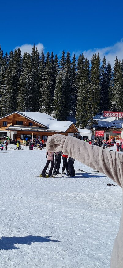 Maskottchen Hopsi steht im Vordergrund und zeigt auf die Skipiste, im Hintergrund sind eine Hütte, Bäume und blauer Himmel zu sehen. | © Planai