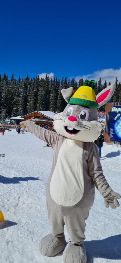 Maskottchen Hopsi steht im Hopsiland im Schnee bei blauem Himmel. | © Planai