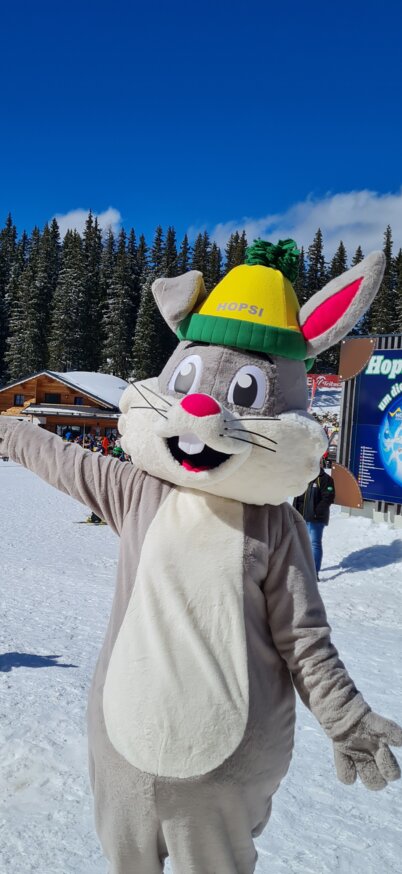 Maskottchen Hopsi steht im Hopsiland im Schnee bei blauem Himmel. | © Planai
