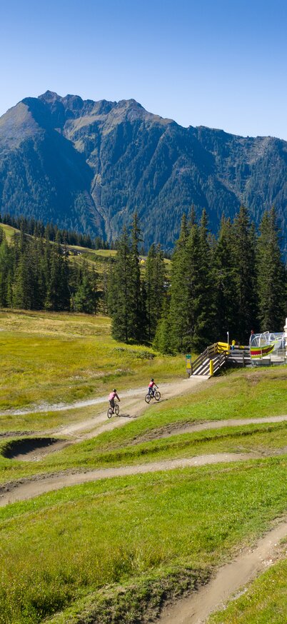 2 Personen mit Mountainbikes fahren in der Bike Area Planai vor Bergkulisse | © Josh Absenger