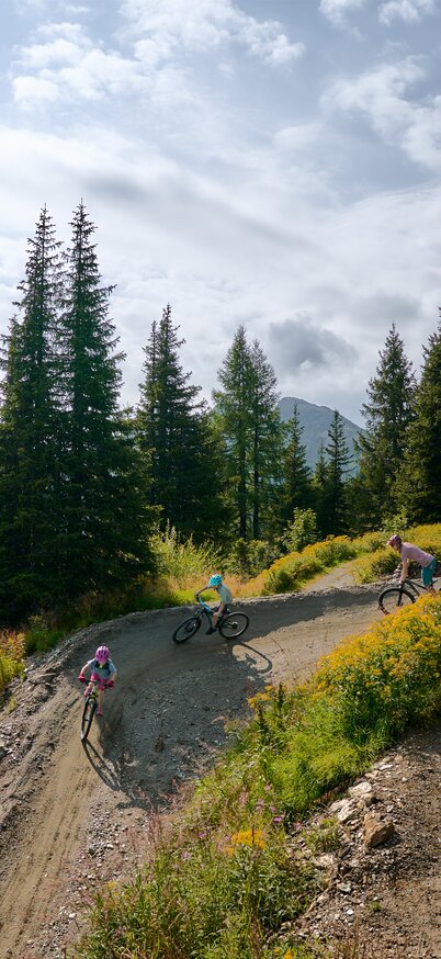 Eine Familie fährt in einer Anliegerkurve auf der Peak Flowline im Bikepark Schladming durch blühende Berglandschaft | © Dietmar Koerbler